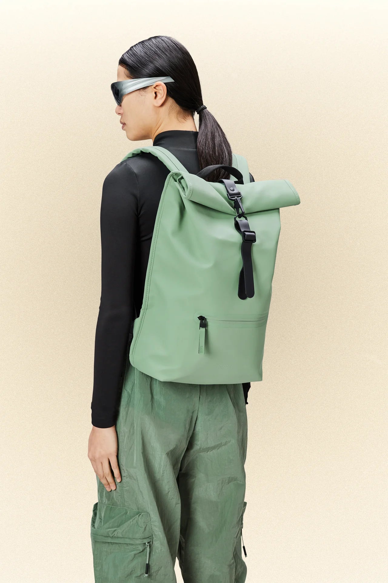 A woman wearing green pants and a Rains Rolltop Rucksack with a laptop pocket.