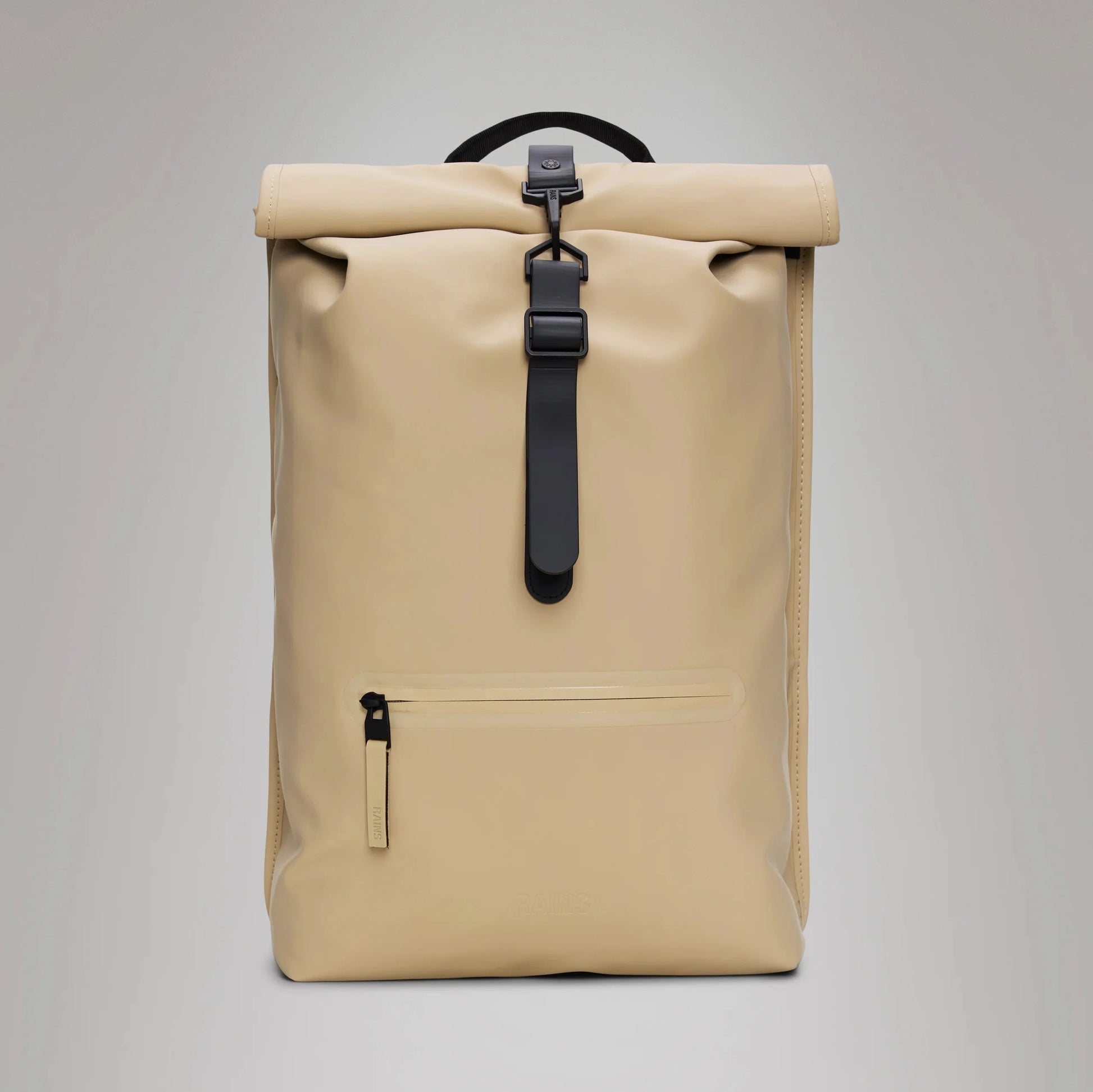 The Rolltop Rucksack by Rains, crafted from waterproof PU fabric, features a beige design with a black front strap, a zipper pocket at the bottom, and a black top handle. It includes a side-access laptop pocket and is styled against a plain light gray background.