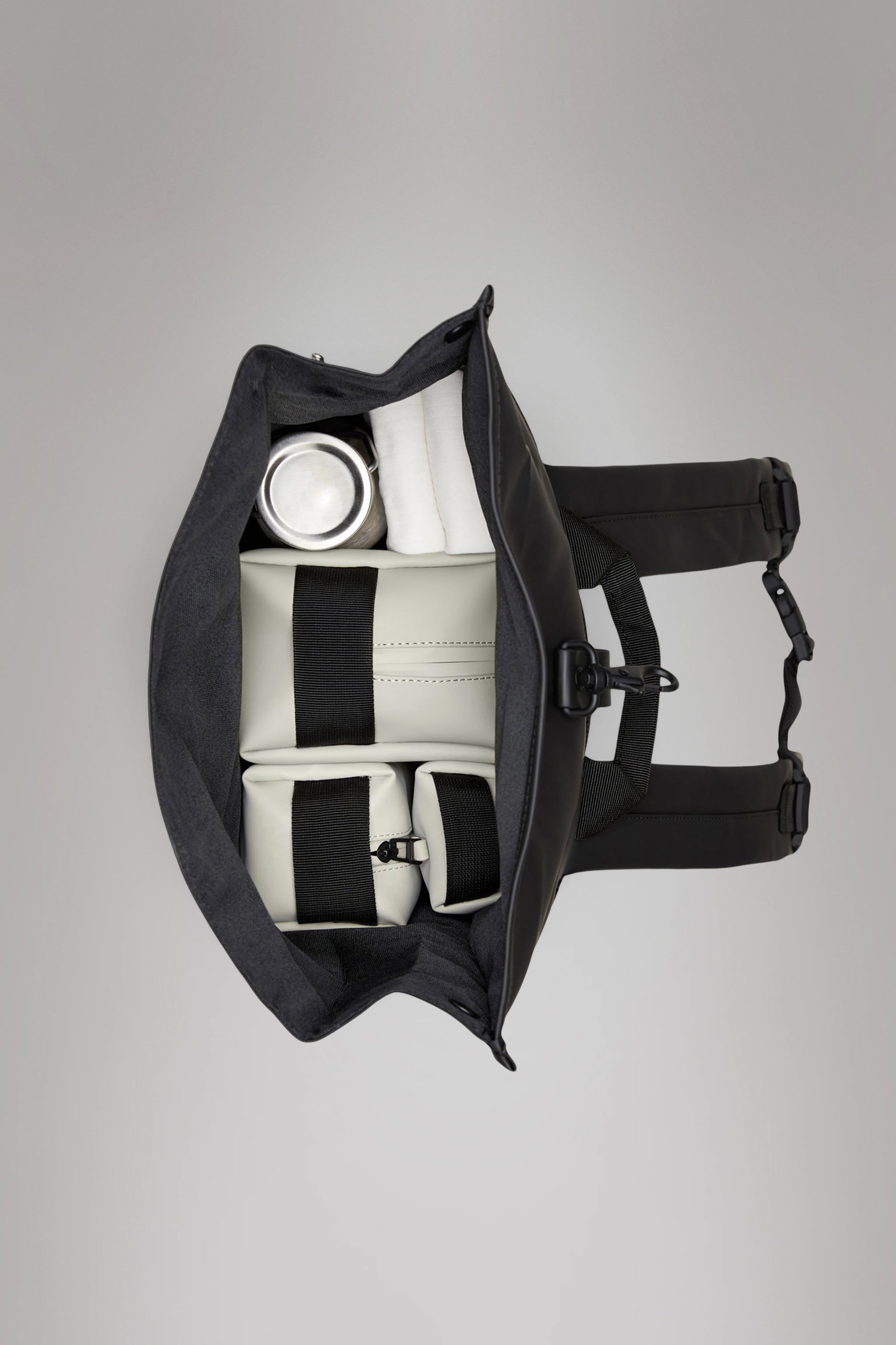 Aerial view of an open black Rains Rolltop Rucksack showcasing a stainless steel water bottle, two white pouches, and a folded white item secured with straps inside. The waterproof PU fabric ensures your essentials stay dry.