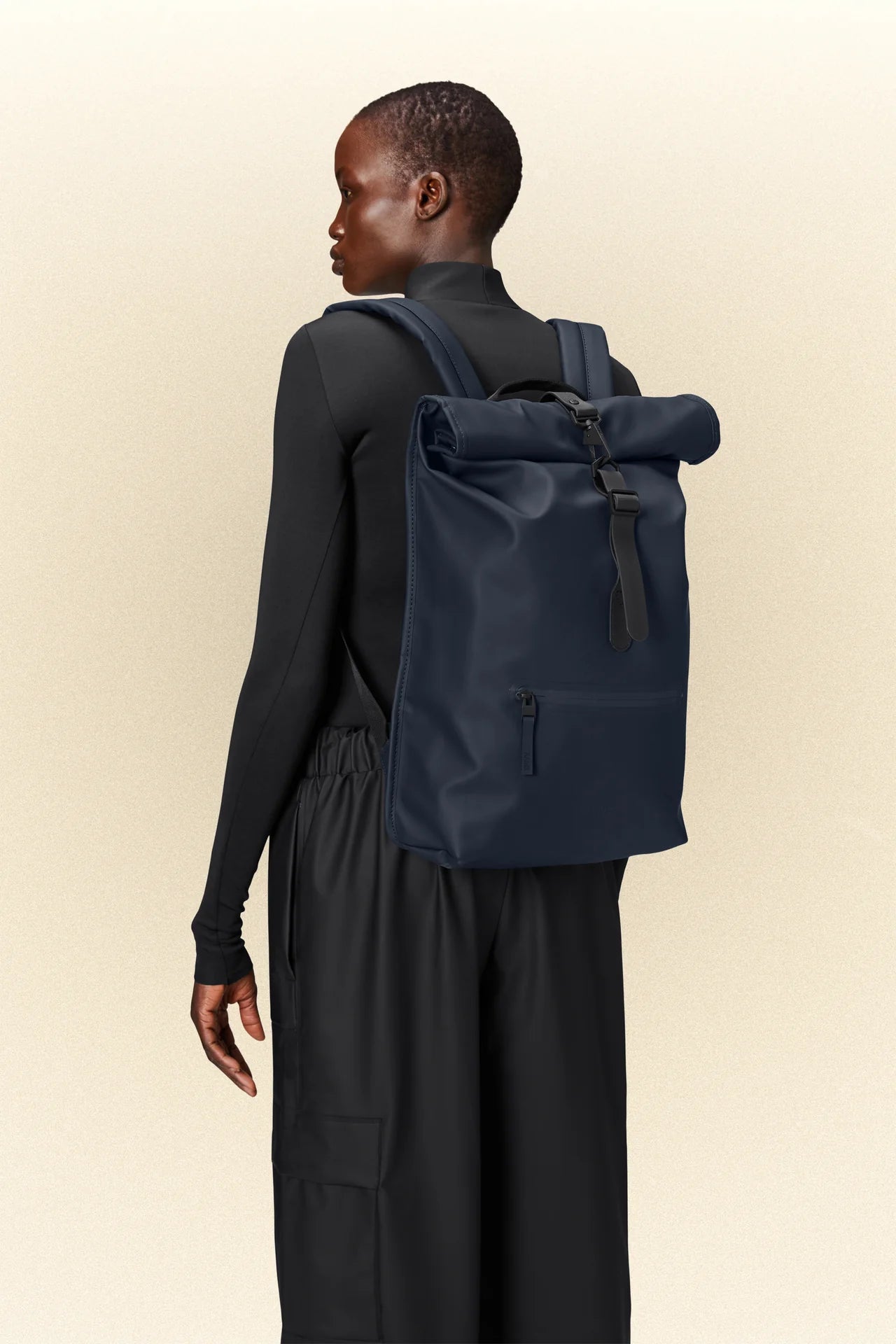 The back view of a woman wearing black pants and a waterproof Rains Rolltop Rucksack with a laptop pocket.