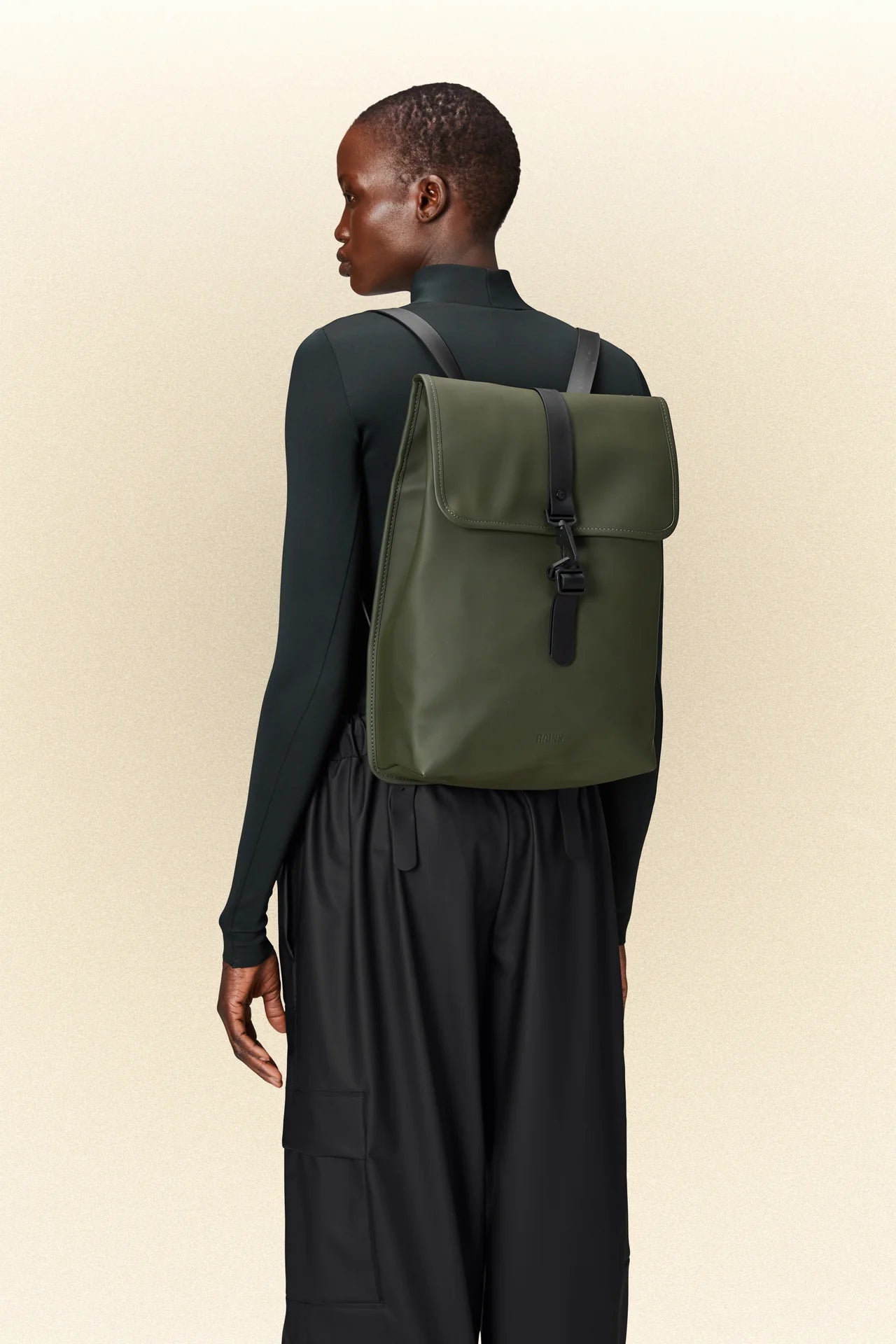 The back view of a woman wearing black pants and a green Rains rucksack with a laptop pocket.
