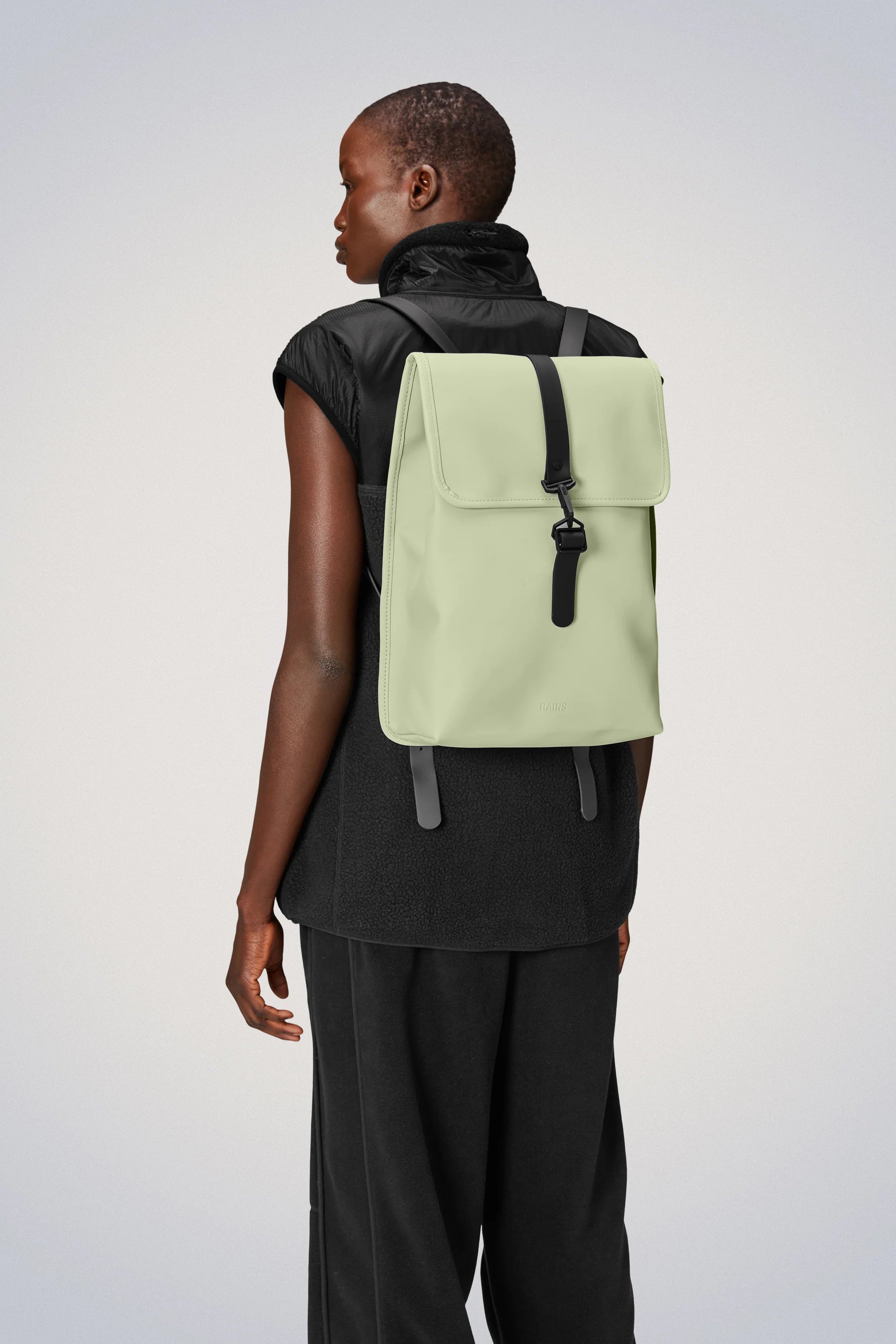 The back view of a woman wearing a green Rains Rucksack. This stylish backpack, made from waterproof fabric, combines practicality with style to meet all your needs. Perfect for daily commutes.