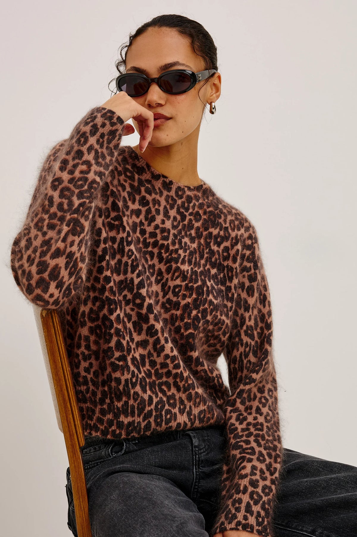 A person wearing black sunglasses, dark jeans, and the Sofia Sweater - Brown Nala by Rails sits on a wooden chair against a plain background.