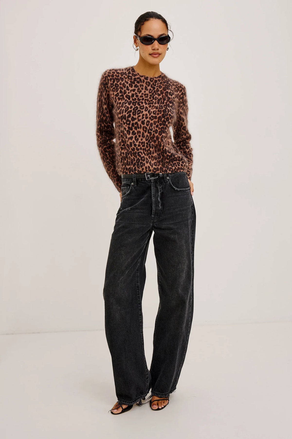 A woman stands against a plain background, wearing sunglasses, loose black jeans, black sandals, and the Rails Sofia Sweater in the Brown Nala leopard print.