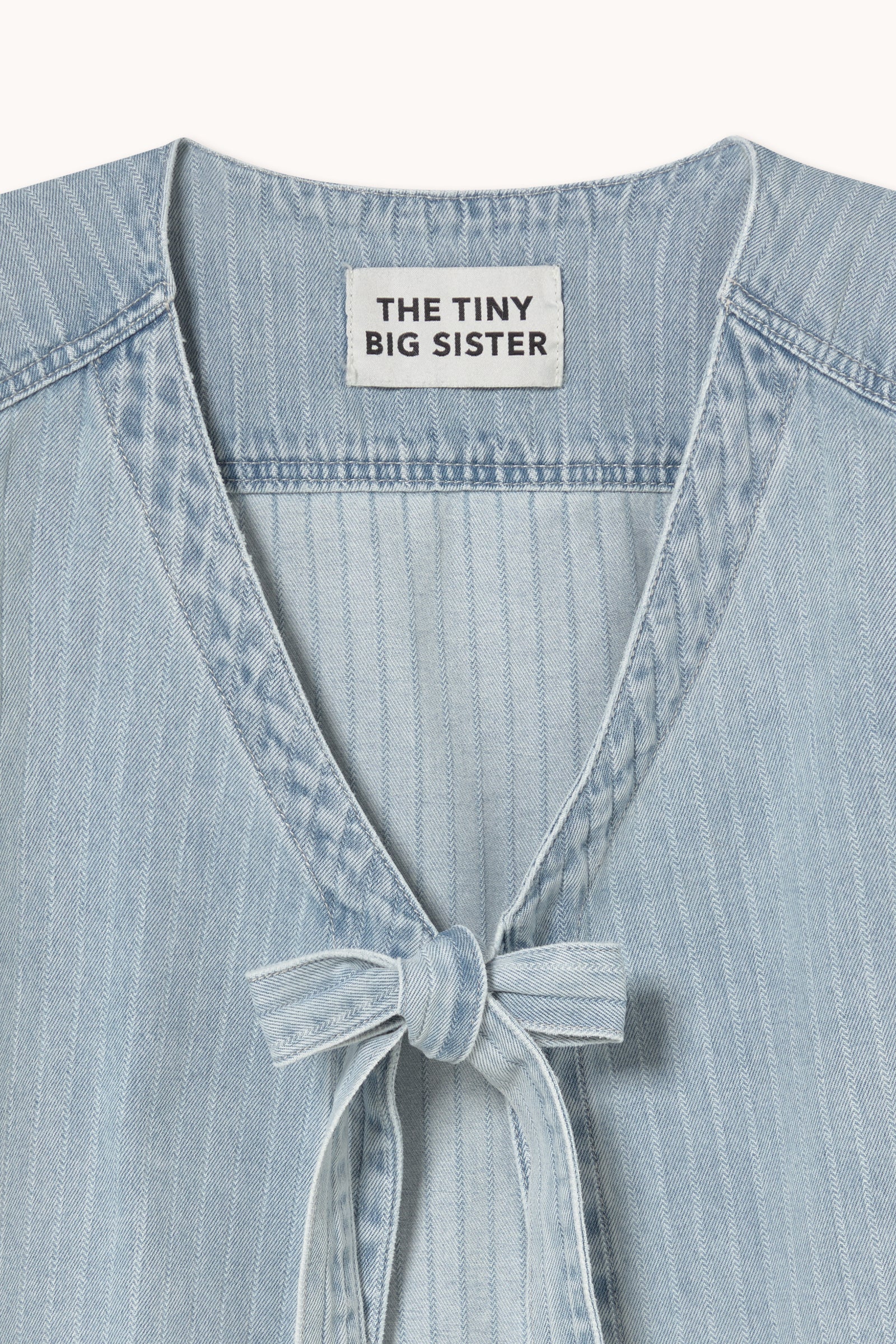 The Denim Tie-Vest in light blue by Tiny Big Sister features a light-wash pinstripe design, a tie-front closure, and an interior label with the brand name.