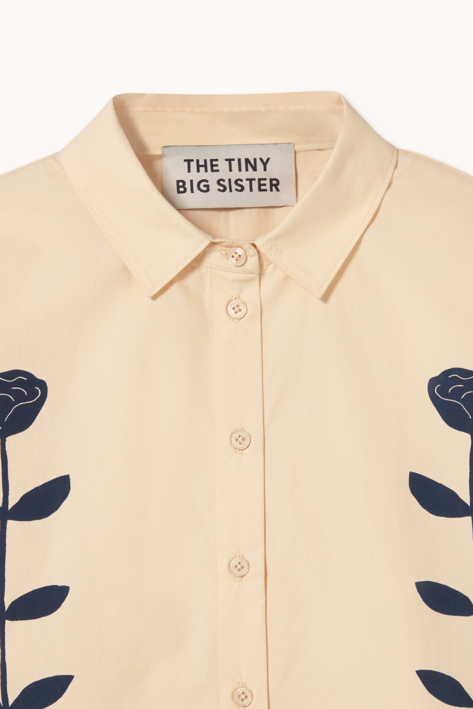 The Flower Shirt - Ivory by Tiny Big Sister is a cream button-up with a collar, black side floral motifs, an oversized boxy fit, and a "THE TINY BIG SISTER" label for effortless style.