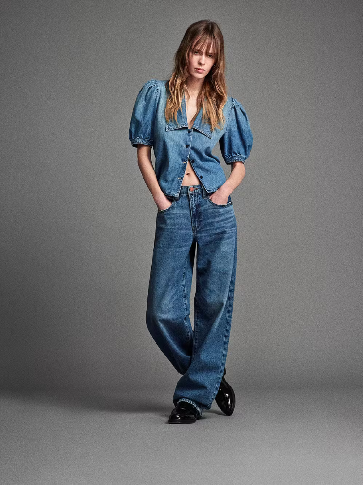 A model stands against a gray backdrop, wearing a denim blouse with puffy sleeves, FRAME Long Barrel Leg jeans in Caramia made from sustainable denim, and black shoes. Hands in pockets, she looks down.