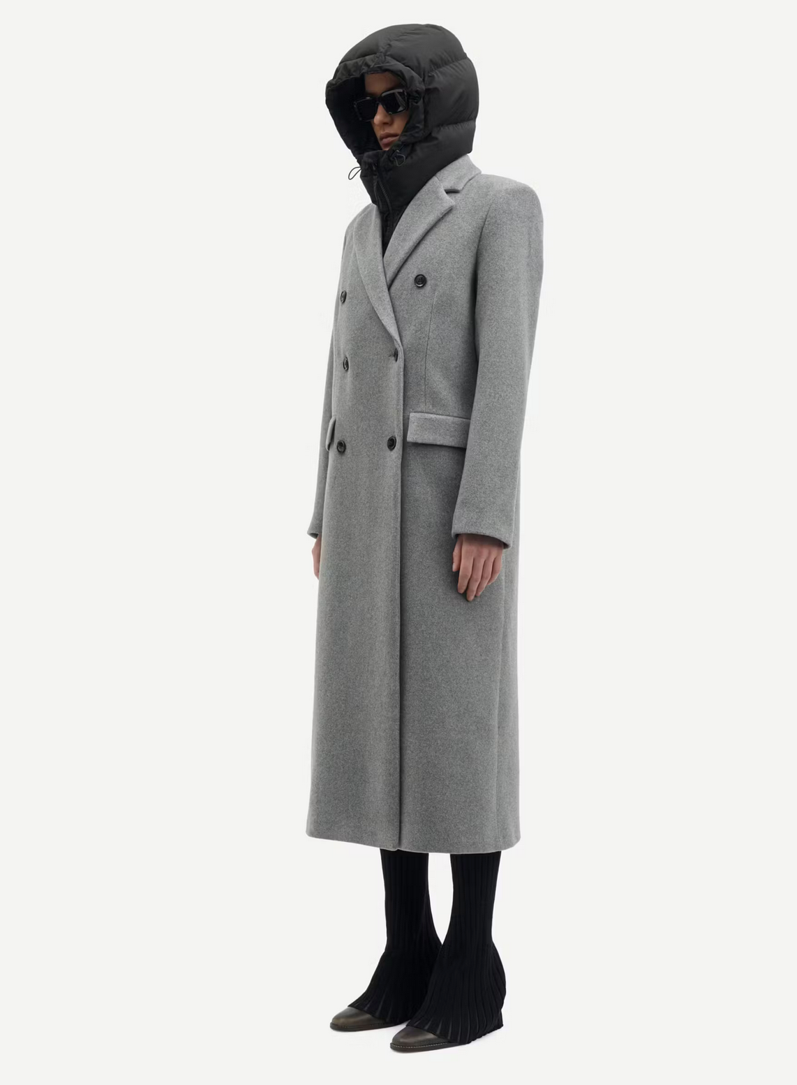 A person is wearing an elegant long "Falcon Coat - Grey Melange" by Samsøe Samsøe, a black hooded jacket, black tights, black sunglasses, and black shoes.