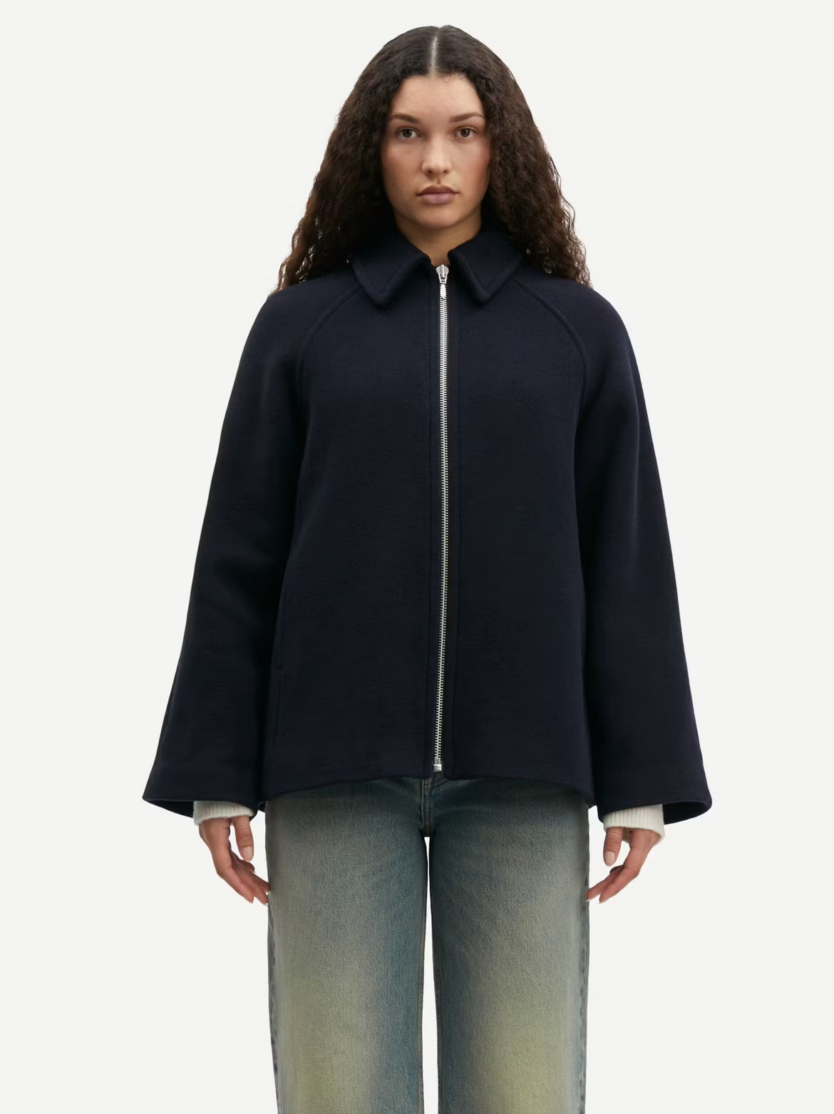 A person with long curly hair is wearing the Alma Jacket in Outer Space by Samsøe Samsøe, made from recycled wool, over a white top and light blue jeans, standing against a plain white background.