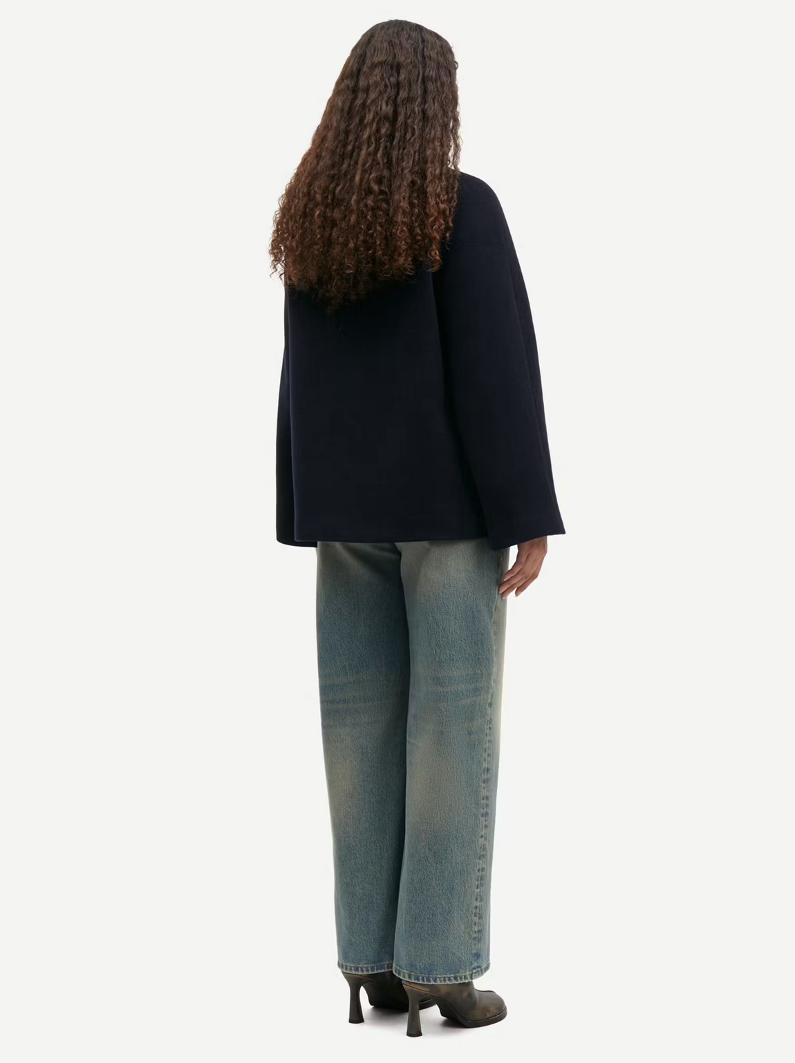 A person with long curly hair is standing with their back facing the camera, wearing a dark long-sleeved top, light blue wide-leg jeans, and dark heeled shoes. Draped over one arm is the stylish Alma Jacket - Outer Space by Samsøe Samsøe, made from recycled wool.