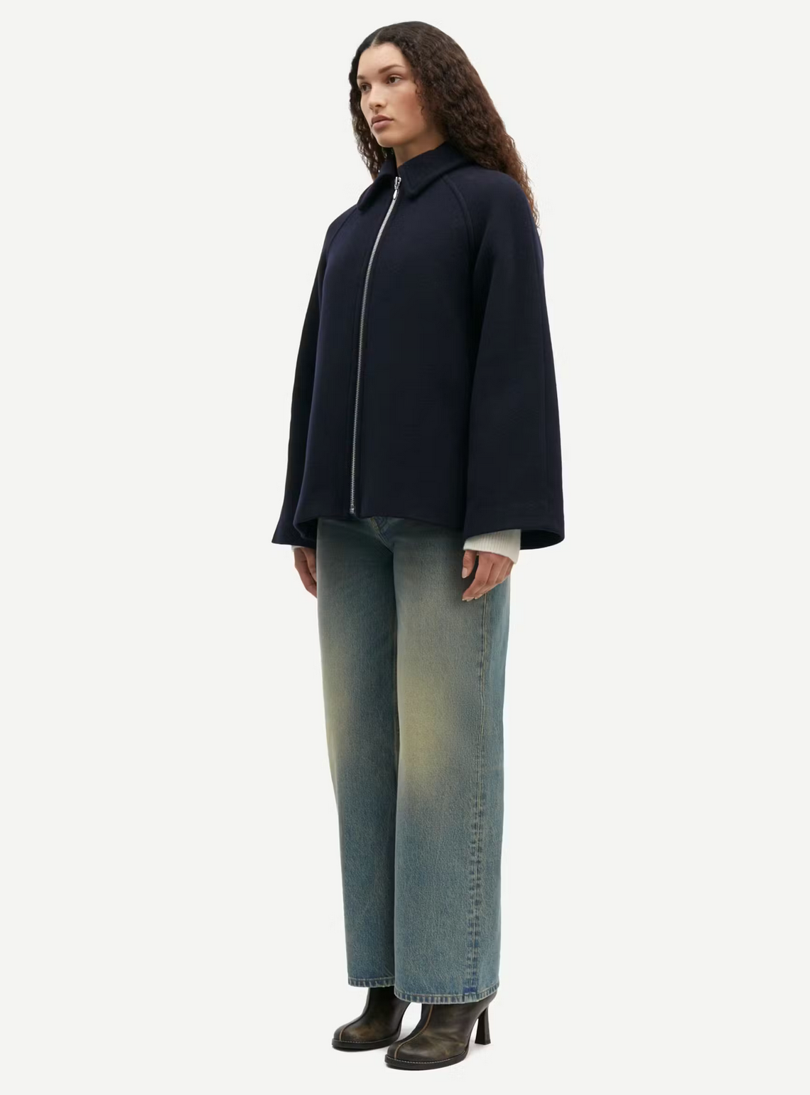 A person stands wearing a Samsøe Samsøe Alma Jacket - Outer Space paired with light blue jeans and black heeled shoes against a plain white background.