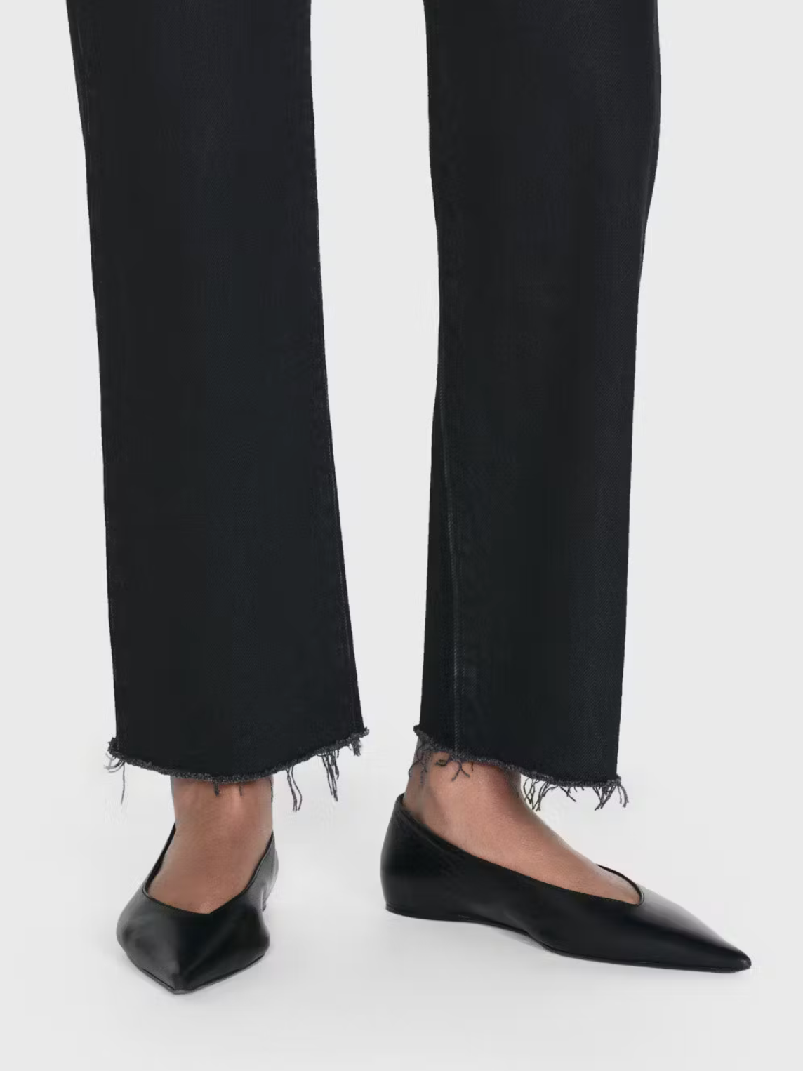 A close-up of a person wearing black Le Jane Crop Raw Fray jeans by Frame and black pointed-toe flats.