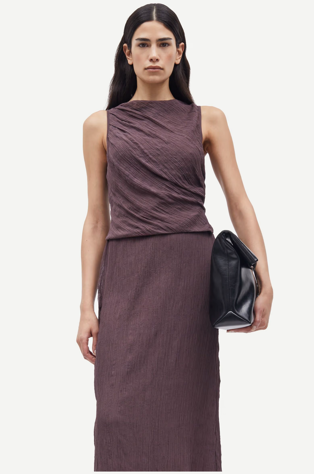 A woman with long dark hair wears the Samsøe Samsøe Savira Top in raisin, featuring a textured sleeveless design made from recycled polyester, and holds a large black clutch.