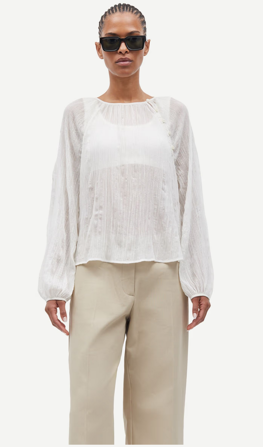 A person in the Savira Blouse - Egret by Samsøe Samsøe, sunglasses, and beige pants stands against a plain white background; the sheer blouse is made from recycled polyester and features long sleeves.