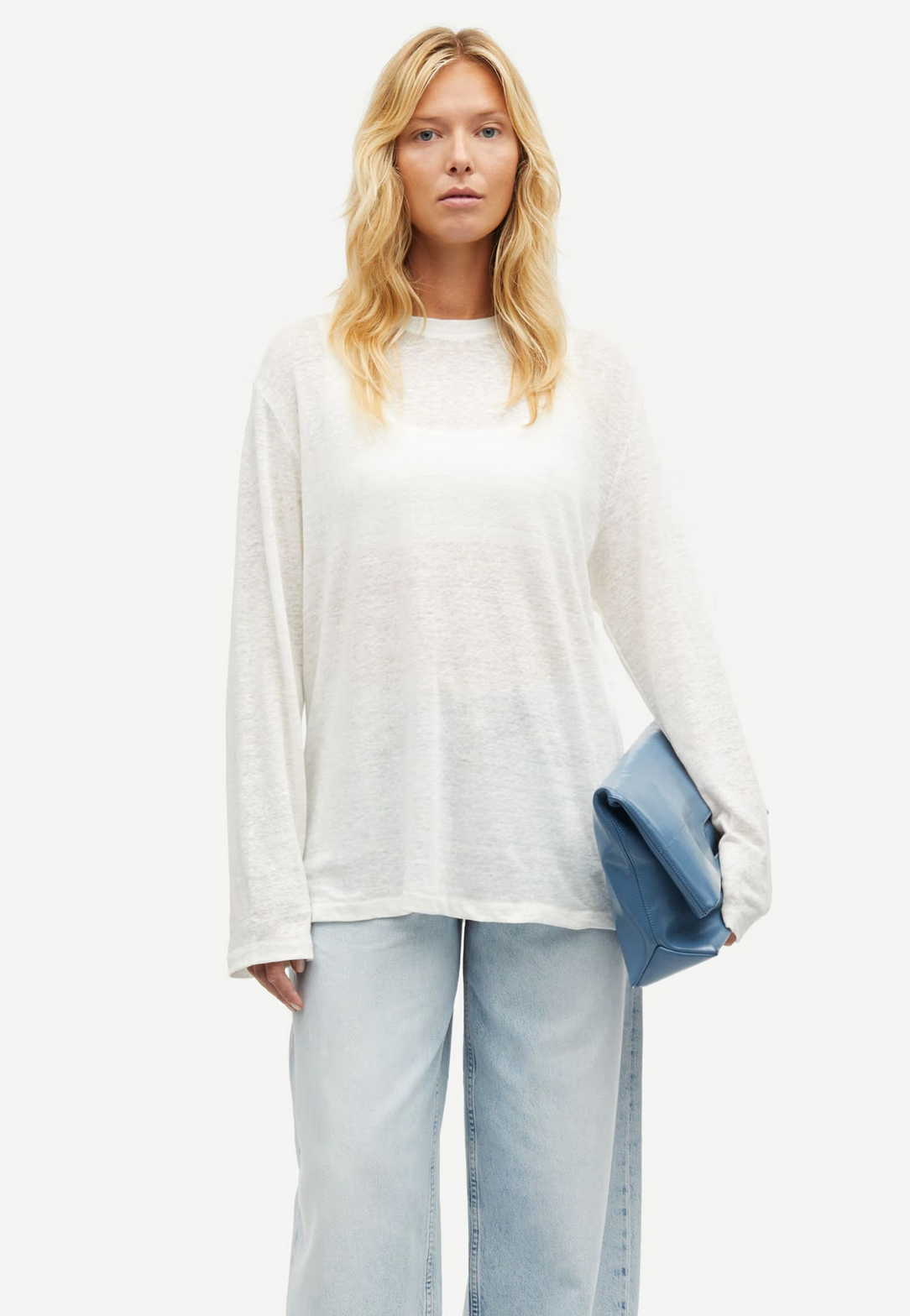 A person with long blonde hair wears the Sadove Long Sleeve Top in Egret by Samsøe Samsøe and light blue jeans, holding a blue clutch against a plain white backdrop.