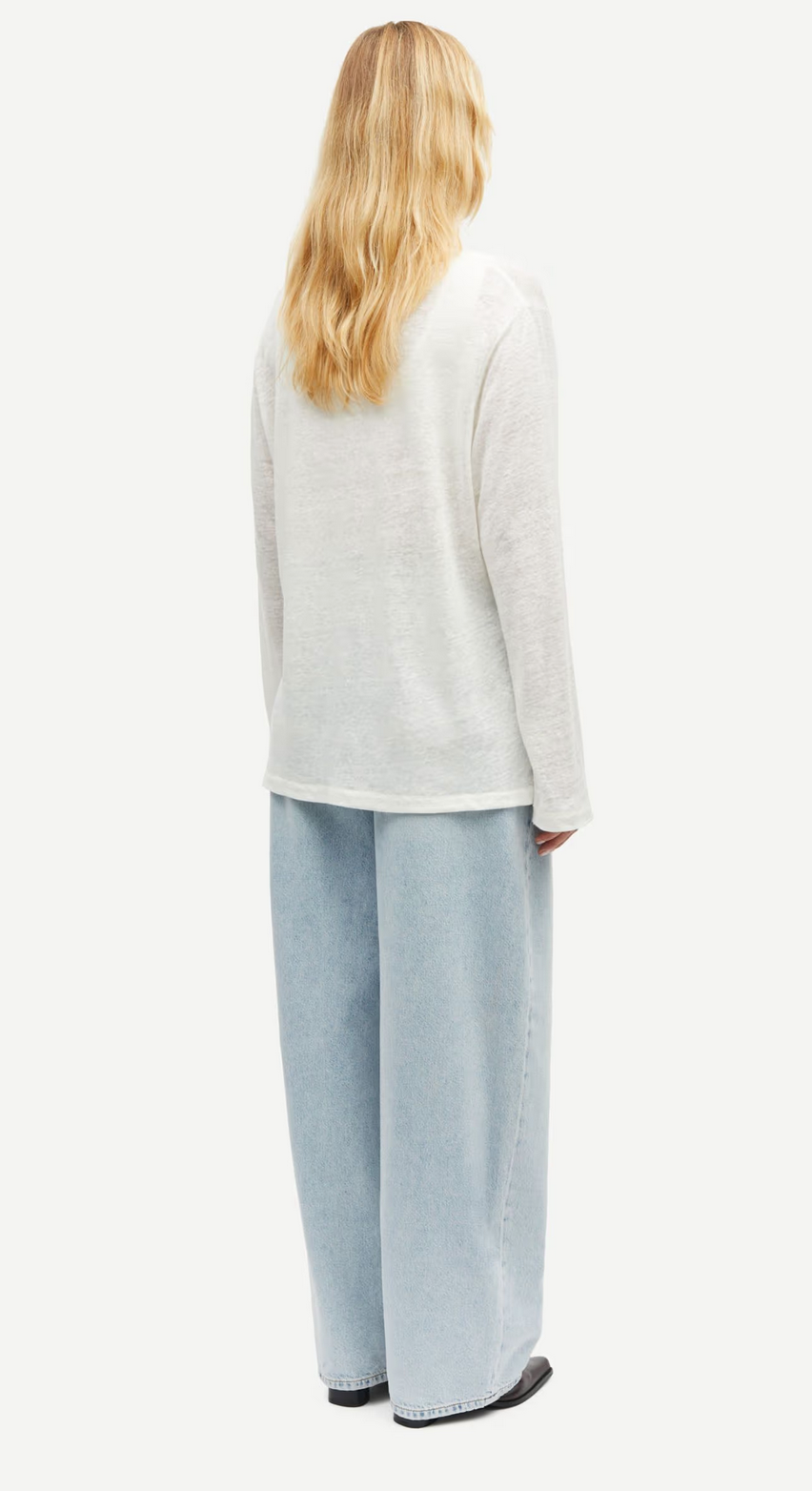 Person with long blond hair wearing a Samsøe Samsøe Sadove Long Sleeve Top in Egret, light blue wide-leg jeans crafted from organic linen, and black shoes, facing away.