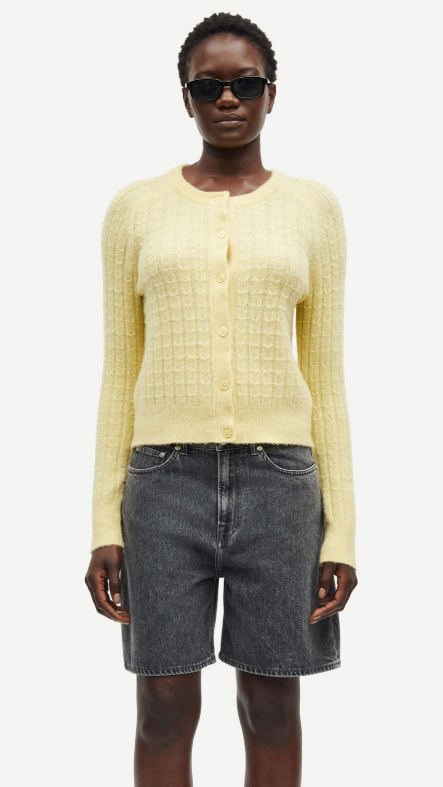 Someone wearing sunglasses and the Samsøe Samsøe Sabarbara Cardigan in Dusty Yellow, paired with gray denim shorts, stands stylishly against a white background.