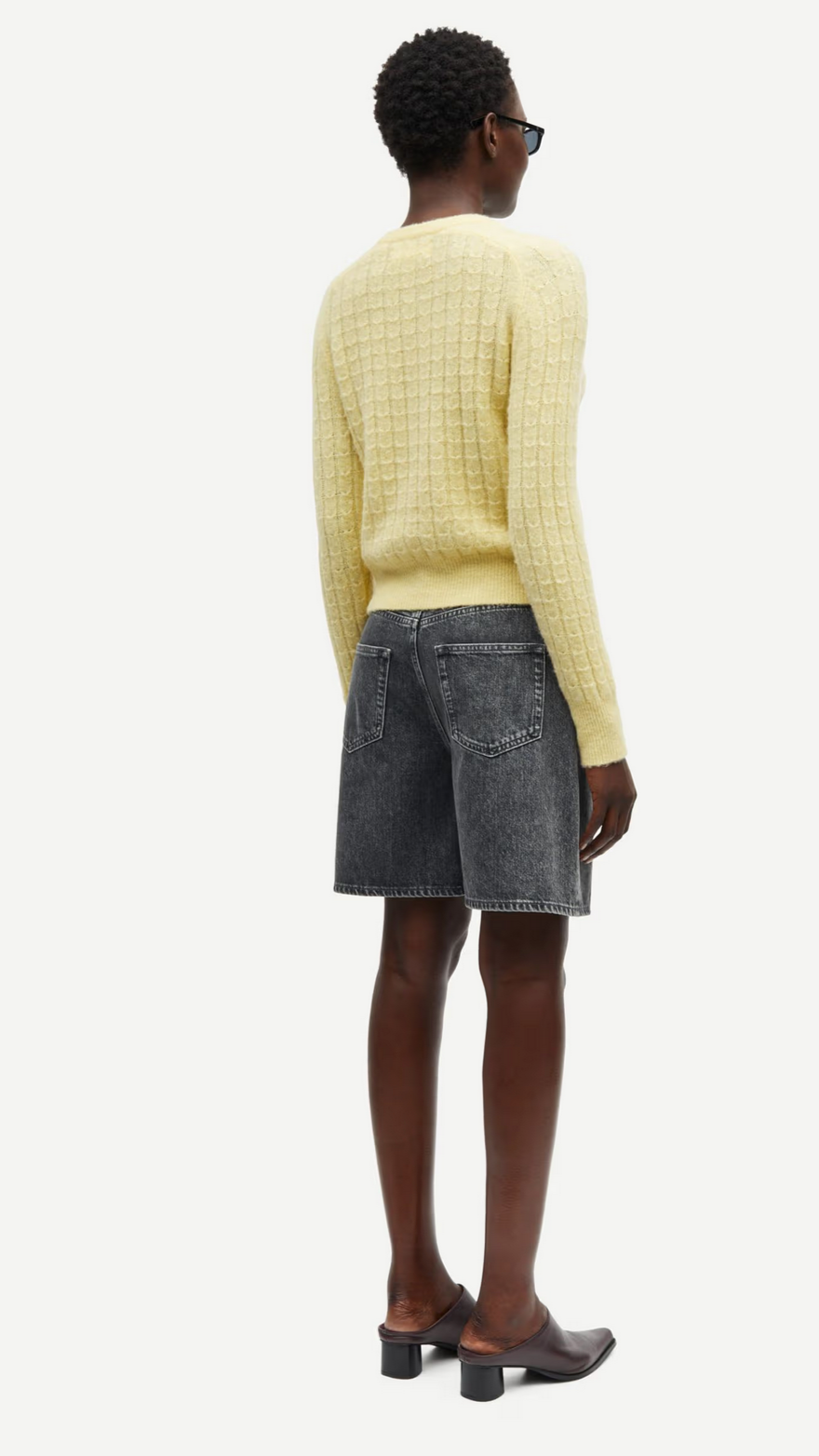 A person wears the Sabarbara Cardigan in Dusty Yellow by Samsøe Samsøe over denim shorts and heeled boots, with their back to the camera.