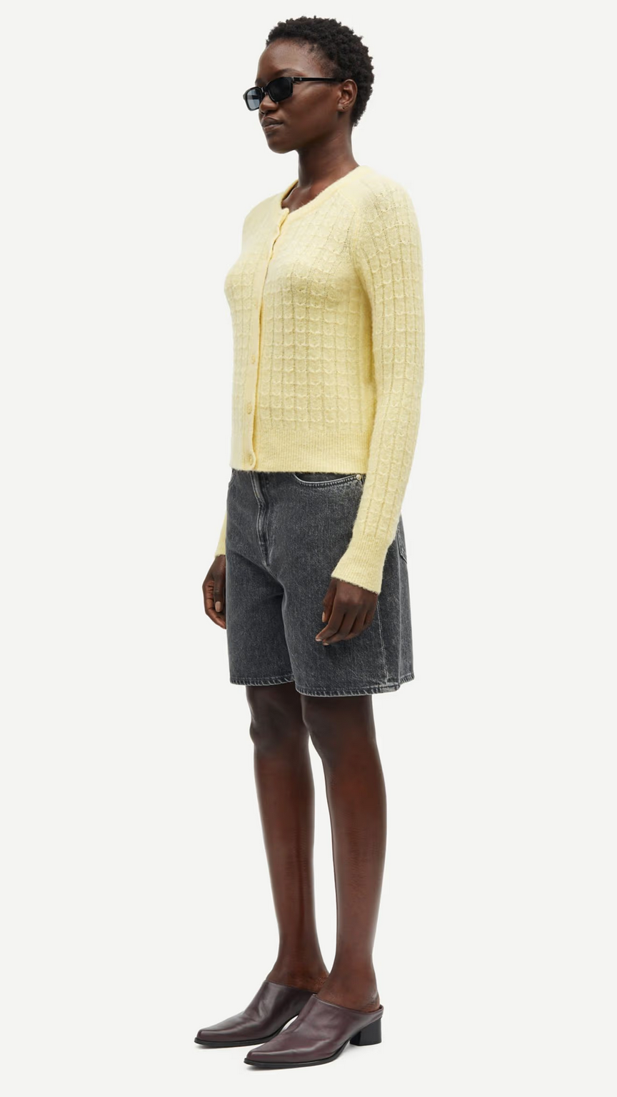 A woman wearing the Samsøe Samsøe Sabarbara Cardigan in Dusty Yellow, paired with black shorts.