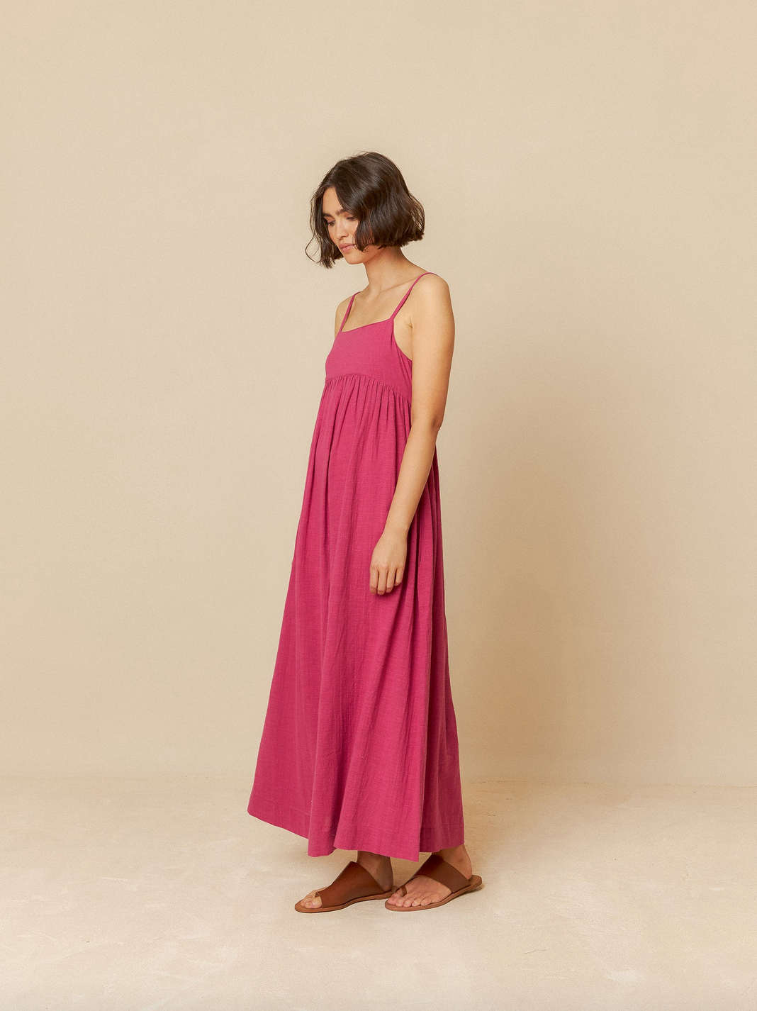 A woman in the Indi & Cold Maxi Dress with Straps - Rose stands sideways against a beige background, looking down. She has short brown hair and wears brown sandals.