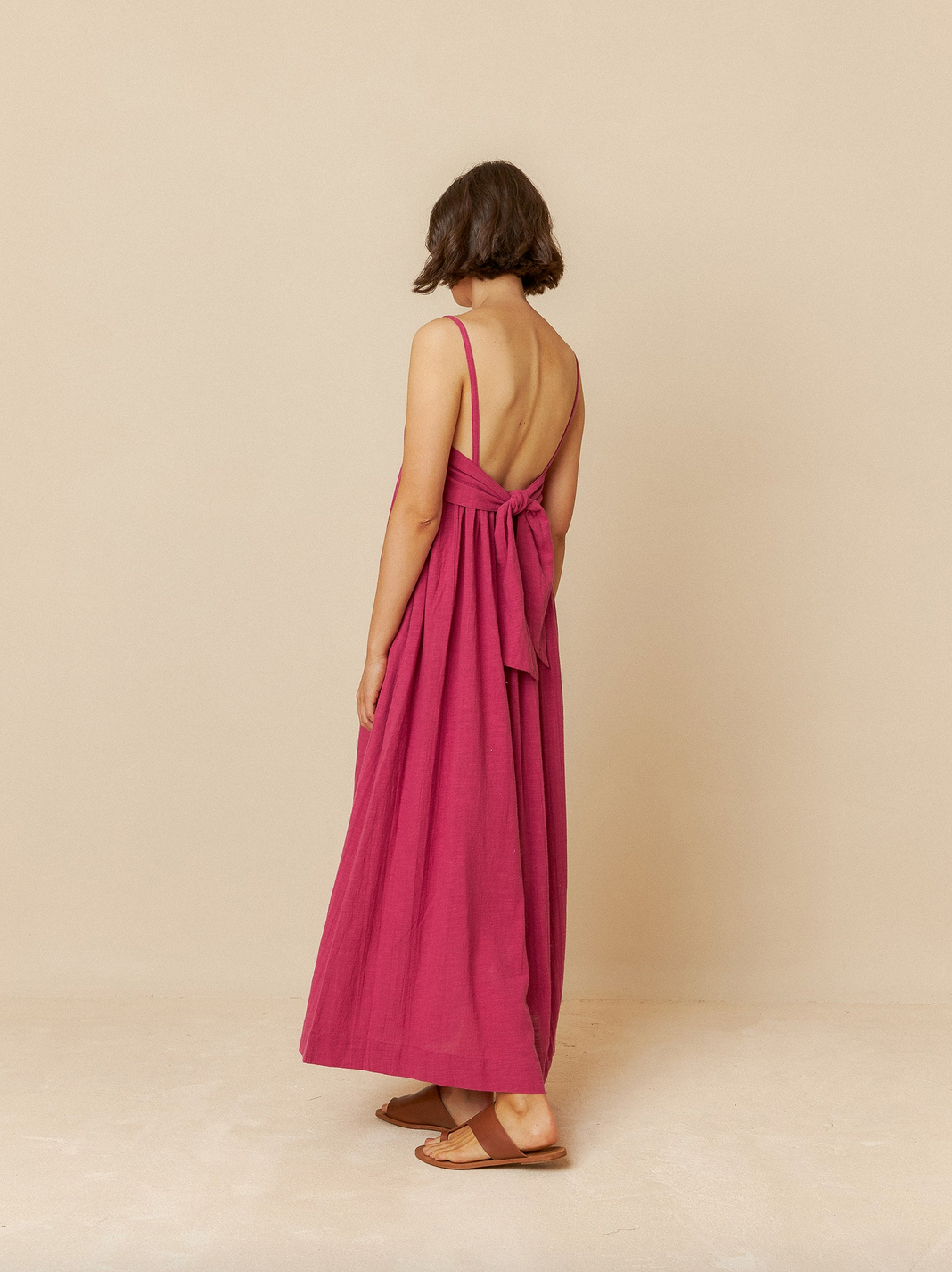 An individual in an Indi & Cold Maxi Dress with Straps - Rose, featuring a bow at the back, stands against a plain backdrop.