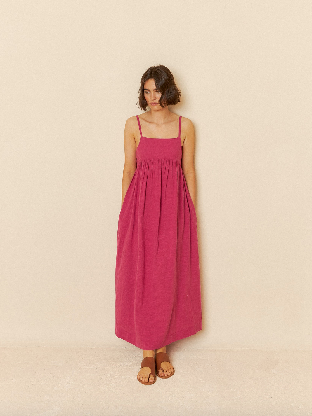 A person in a Maxi Dress with Straps - Rose by Indi & Cold stands against a plain beige background, wearing brown sandals.