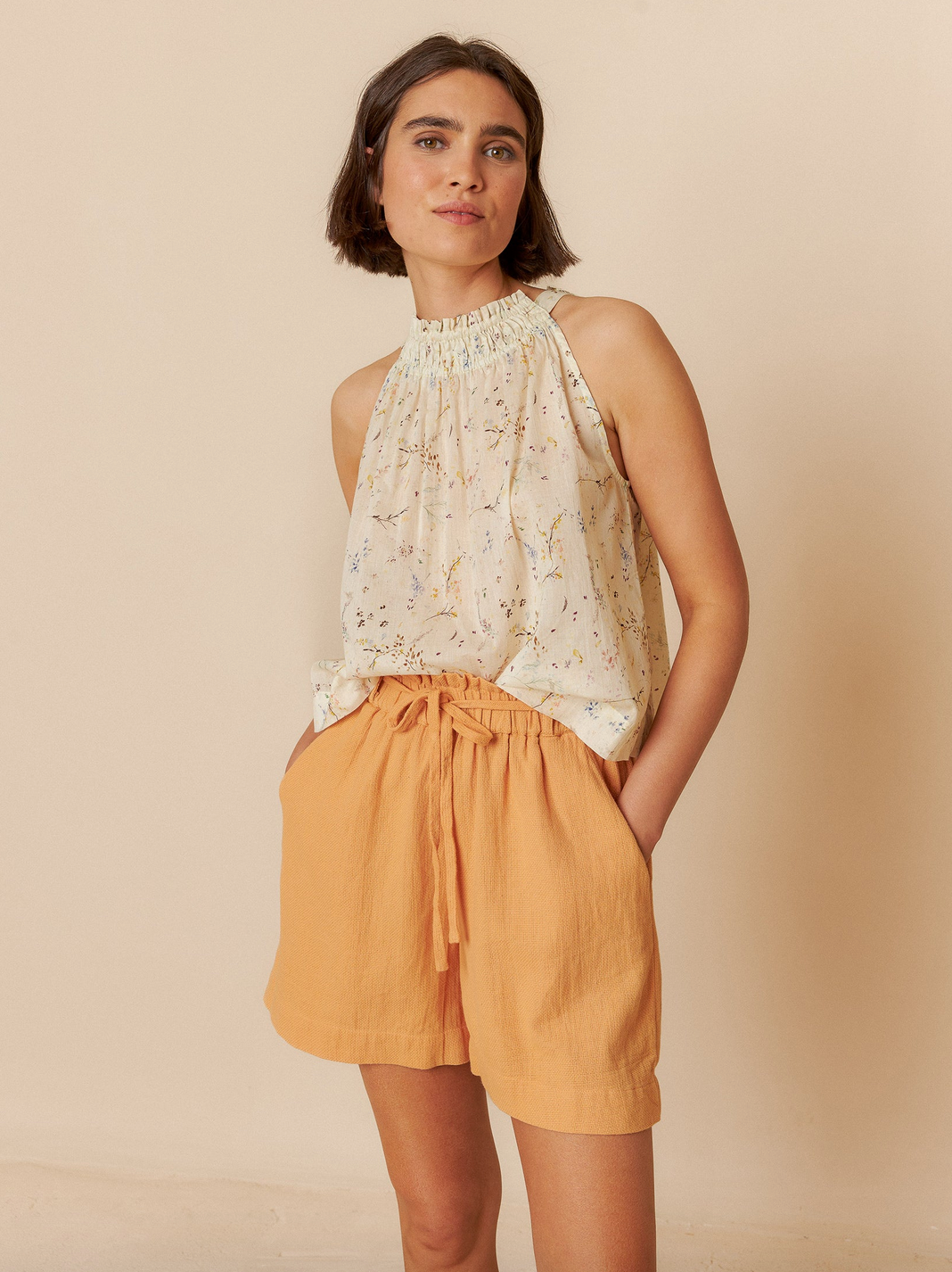 A person poses against a neutral background wearing the Halter Collar Top - Vanilla by Indi & Cold and light orange shorts, hands in pockets.