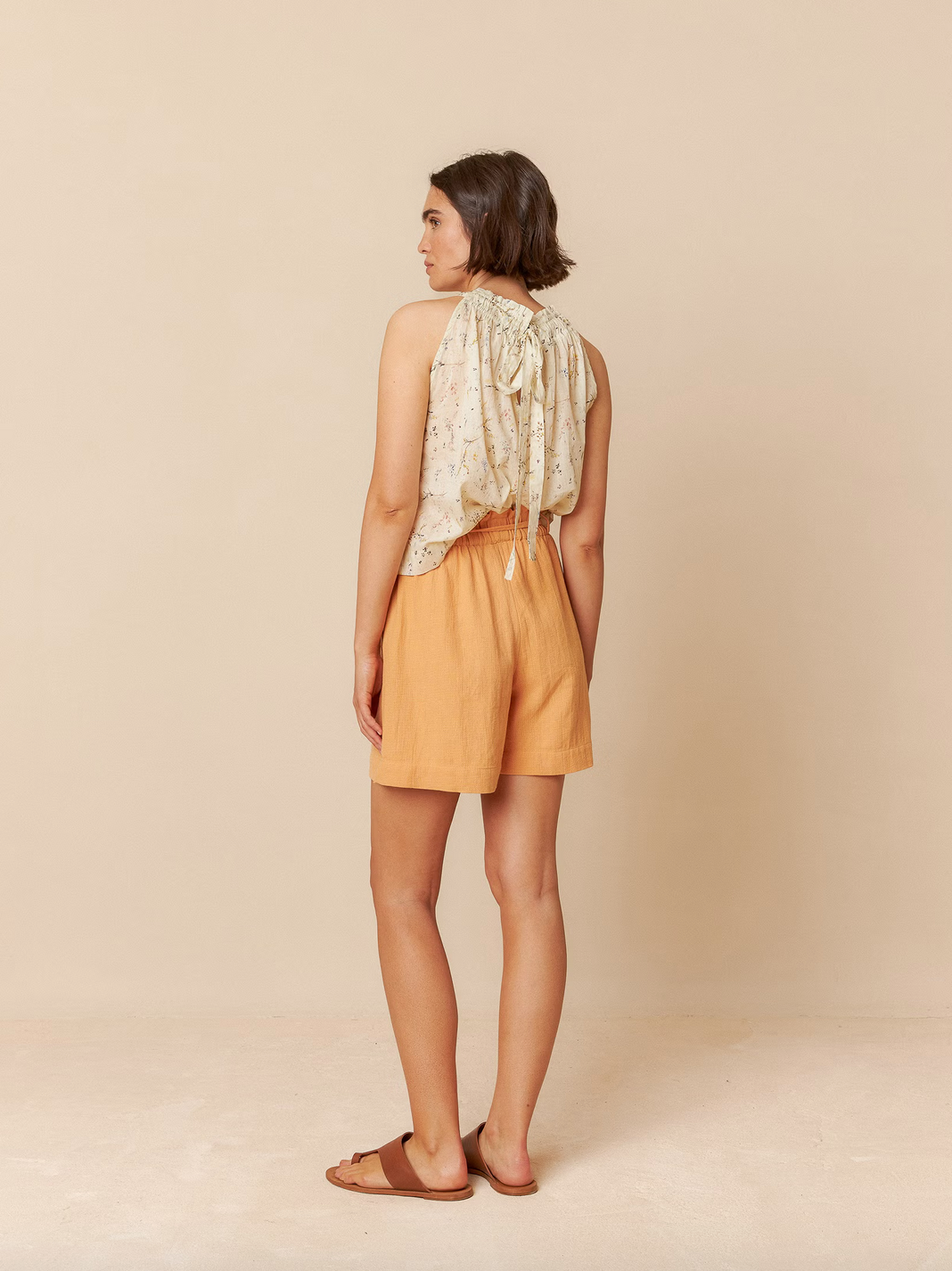 A woman, facing away from a plain beige wall, wears the Indi & Cold Halter Collar Top in vanilla with a floral print, paired with mustard shorts and brown sandals.