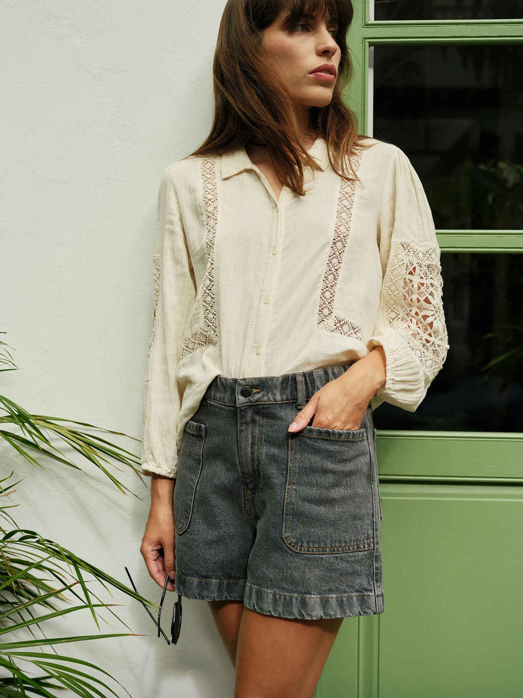 A person in an Indi & Cold Double Gauze Shirt - Ivory and dark denim shorts stands by a green-framed window, holding sunglasses.