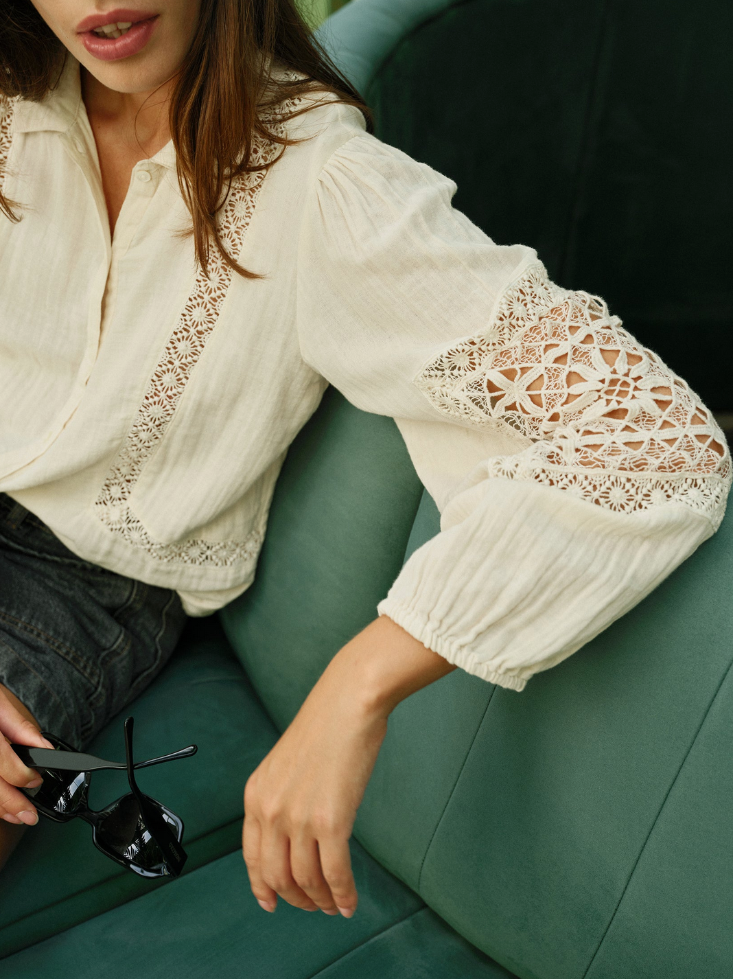 A person in an Indi & Cold Double Gauze Shirt - Ivory sits on a green sofa, holding black sunglasses.