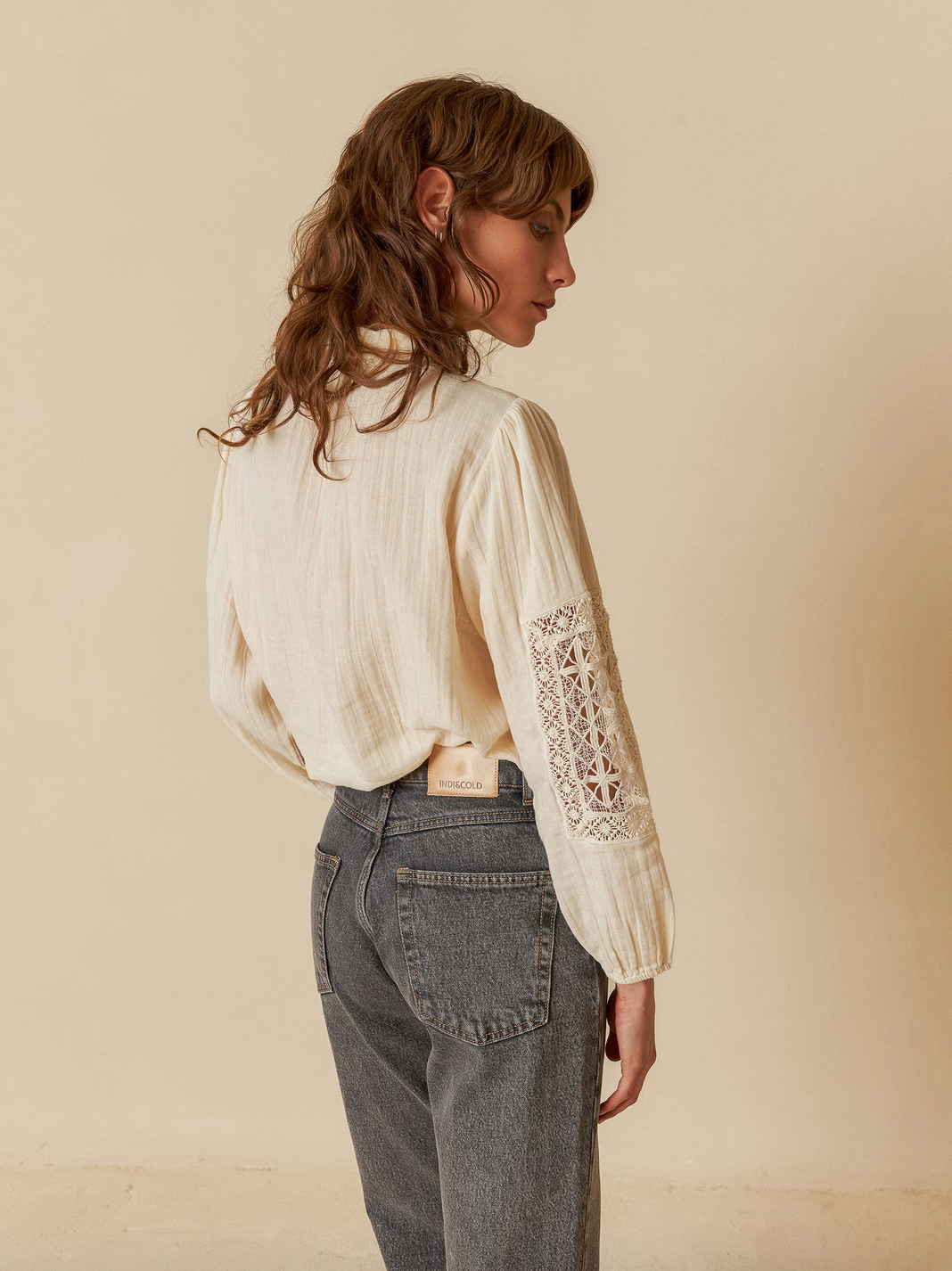 A person in an Indi & Cold Double Gauze Shirt - Ivory with lace sleeve inserts and gray jeans gazes off to the side against a plain beige background.