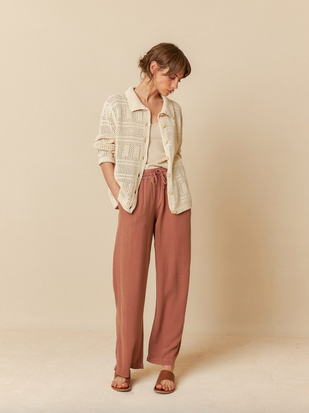 A person in an Indi & Cold Crochet Knit Shirt - Ivory and peach pants stands against a beige background, looking down with hands in pockets.