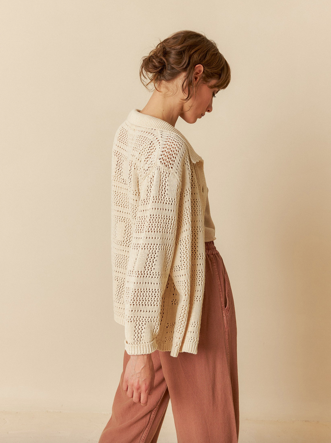 An individual wearing an Indi & Cold Crochet Knit Shirt in Ivory and brown pants stands sideways against a plain beige background.