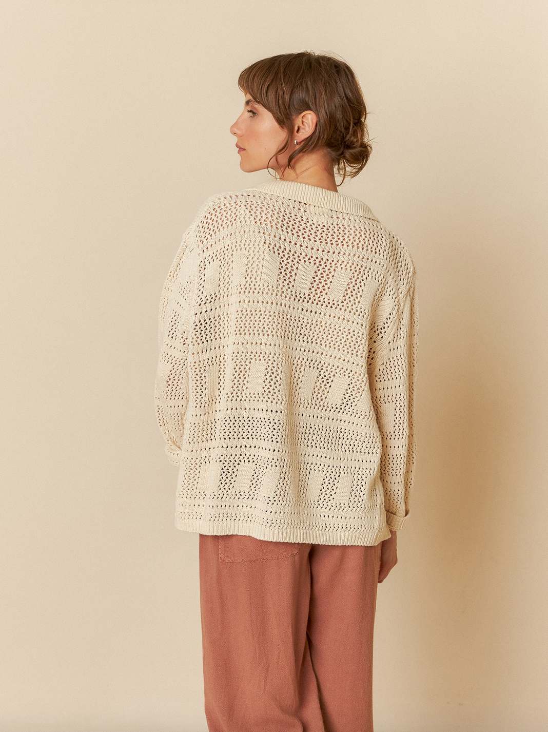 A person, facing away, wears the Crochet Knit Shirt - Ivory by Indi & Cold, crafted from recycled cotton with crochet-effect patterns and geometric motifs, paired with rose-colored pants against a beige backdrop.