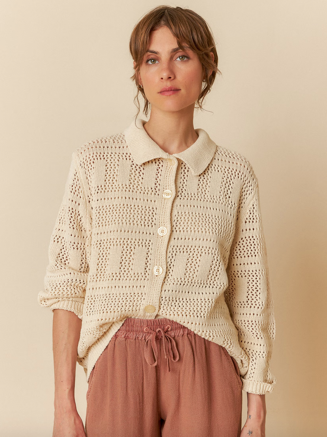 An individual wearing the Indi & Cold Crochet Knit Shirt in ivory and brown pants stands against a beige background, looking forward.