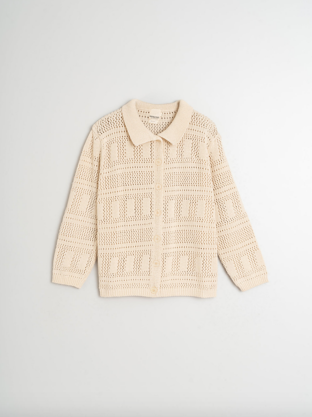 The Crochet Knit Shirt by Indi & Cold is an ivory, long-sleeve cardigan made from crochet-effect knit fabric and recycled cotton, featuring a collared neck and button closure, photographed on a white background.