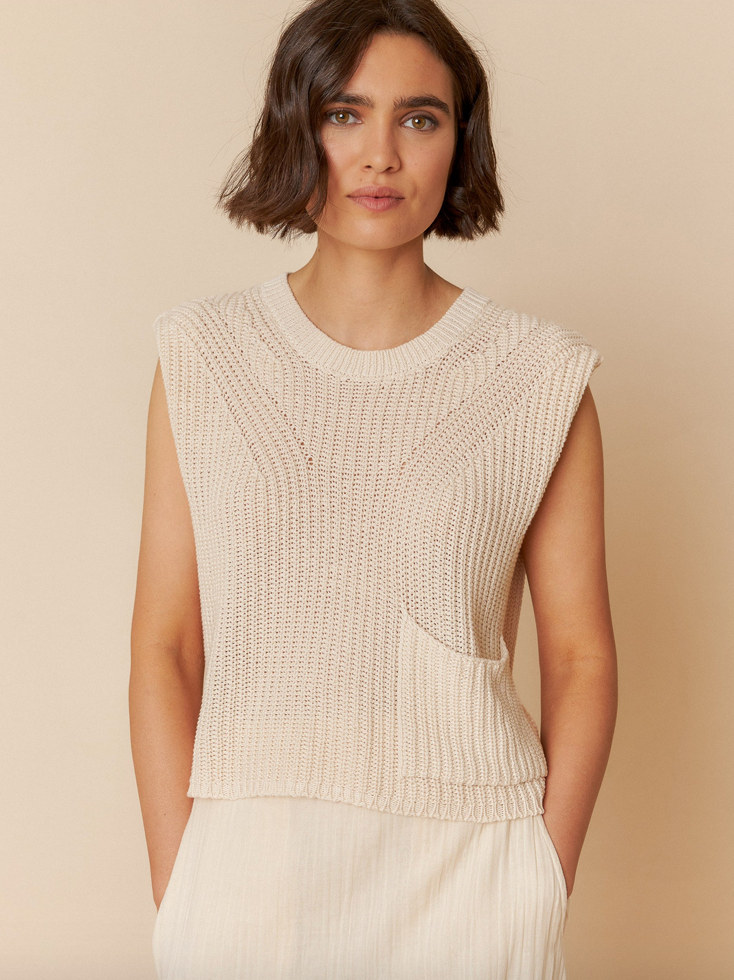 A woman with short brown hair wears the Indi & Cold English Rib Knit Vest in Ivory, which is made from organic cotton and features a stylish pocket, as she stands against a matching beige background.