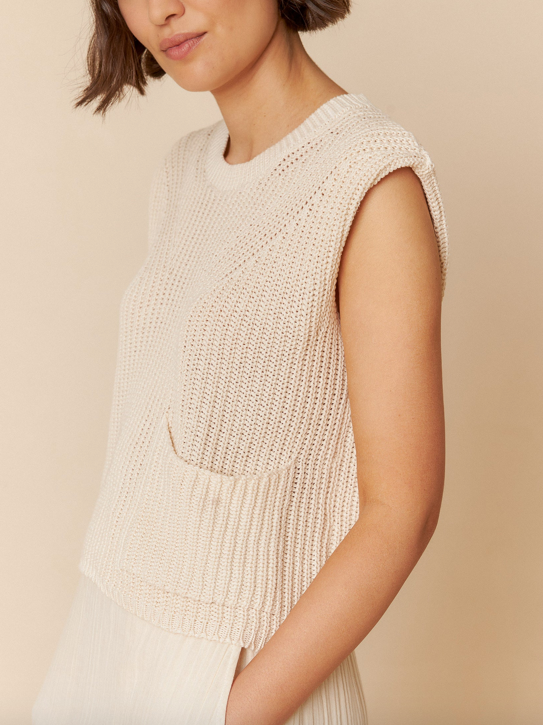 A model elegantly poses against a beige backdrop, donning the versatile Indi & Cold English Rib Knit Vest in ivory paired with an off-white pleated skirt.