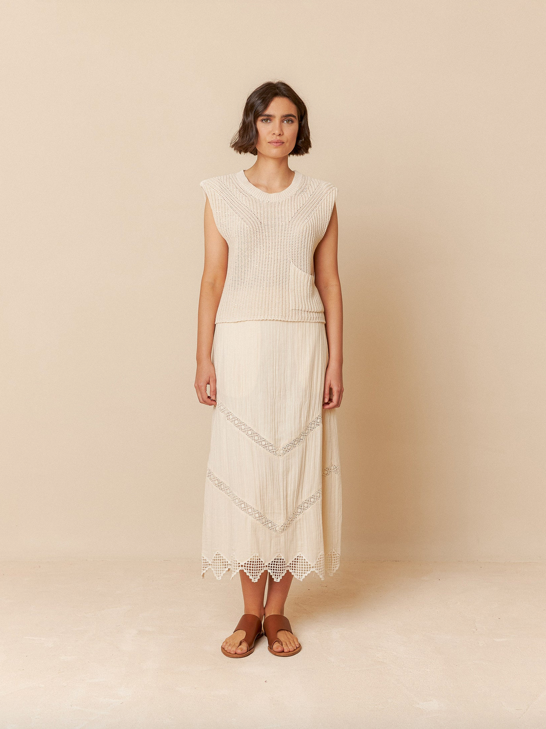 A woman elegantly poses against a beige background in the English Rib Knit Vest - Ivory from Indi & Cold, paired with a matching long skirt, all crafted from organic cotton.