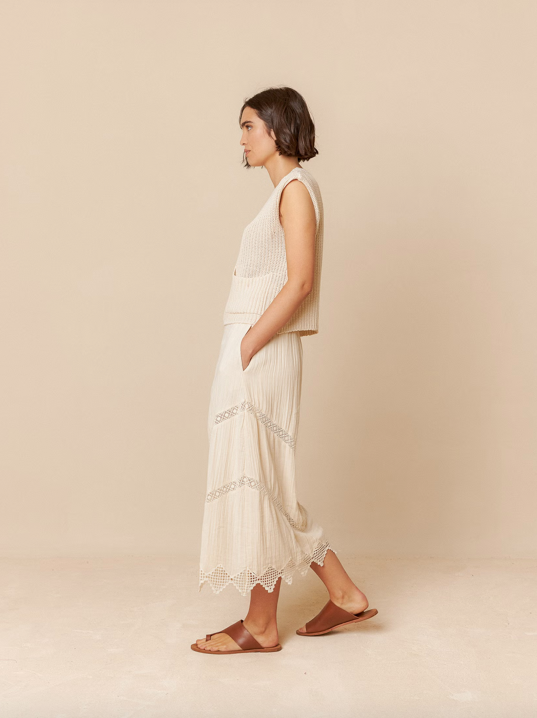 A woman stands in profile against a beige background, wearing an outfit featuring the Indi & Cold English Rib Knit Vest - Ivory and a long skirt with lace details, paired with sandals.