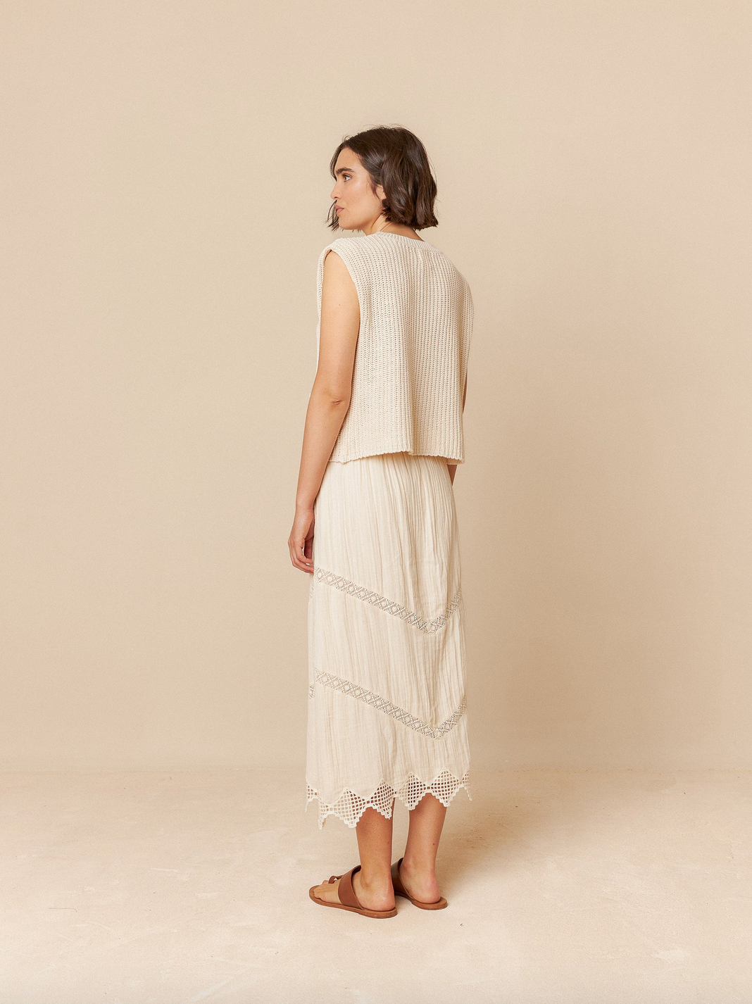 A woman in an Indi & Cold staple, wearing the English Rib Knit Vest in ivory with a long organic cotton skirt, stands barefoot on a beige floor against a soft backdrop, looking left.