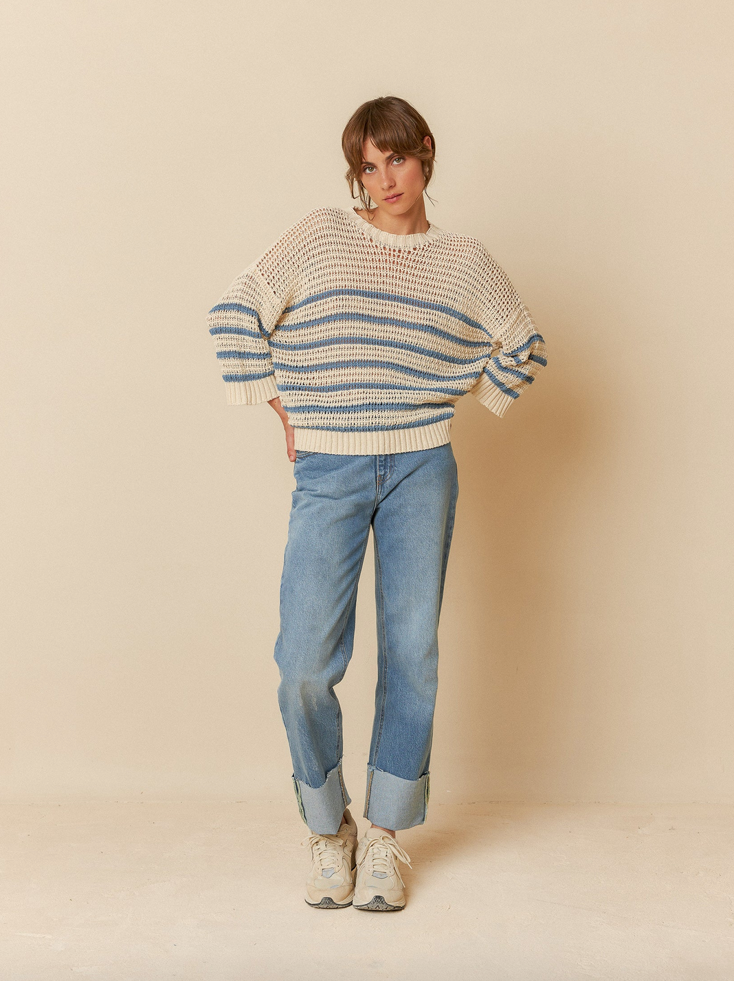 In a neutral setting, an individual showcases the Indi & Cold Dual Stripe Sweater - Indigo in beige and blue stripes, paired with wide-cuffed blue jeans and white sneakers, embodying effortless style.