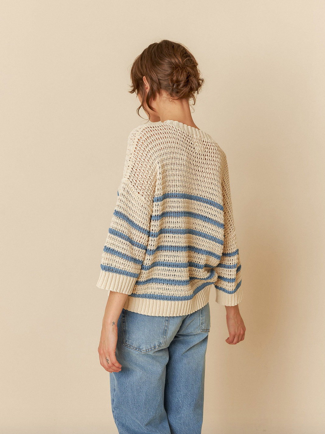 Person in an Indi & Cold Dual Stripe Sweater - Indigo, paired with loose blue jeans, stands facing away against a beige background.