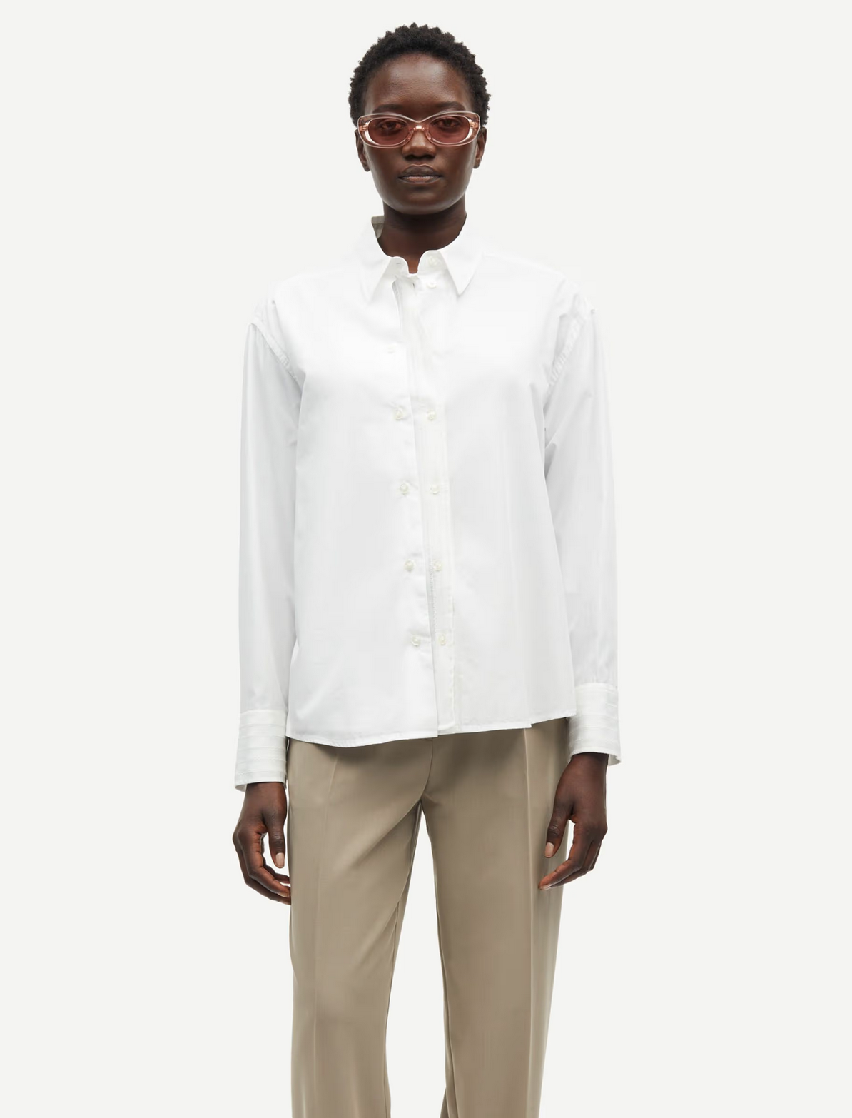 A person in a relaxed fit Salasa Shirt by Samsøe Samsøe, featuring a white button-up long-sleeve design, paired with beige pants and sunglasses, stands against a plain background.