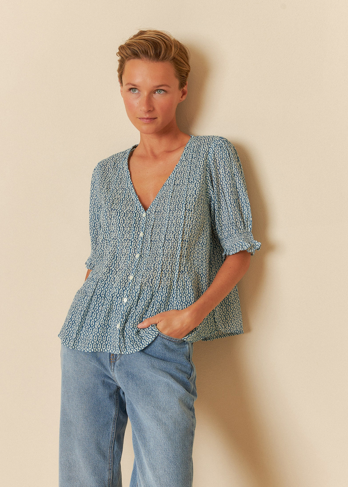 Person with short hair wearing an Indi & Cold Short Sleeve Blouse - Blue, featuring a geometric print and V-neckline, paired with blue jeans against a neutral background.