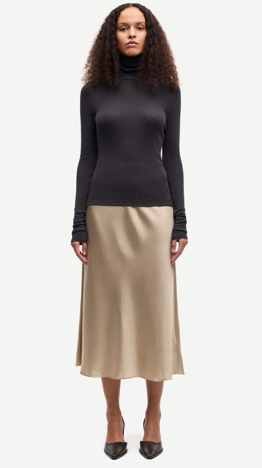 A woman stands facing forward, wearing a black long-sleeve turtleneck top, the Samiley Skirt - Aluminium by Samsøe Samsøe, and black pointed-toe heels.