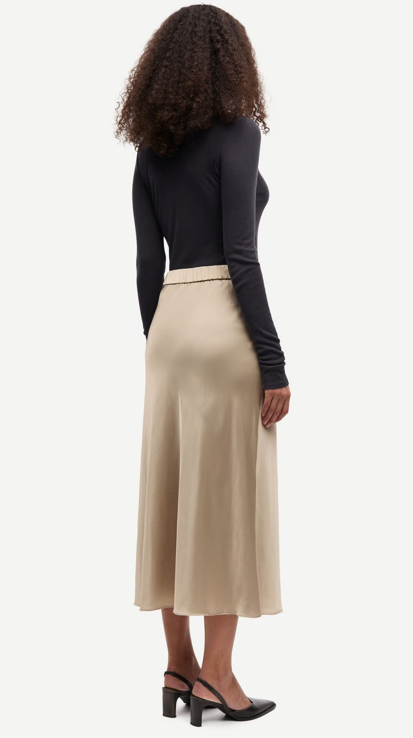 A woman with curly hair faces away, wearing a black long-sleeve top, the Samiley Skirt - Aluminium by Samsøe Samsøe, and black heeled slingback shoes.
