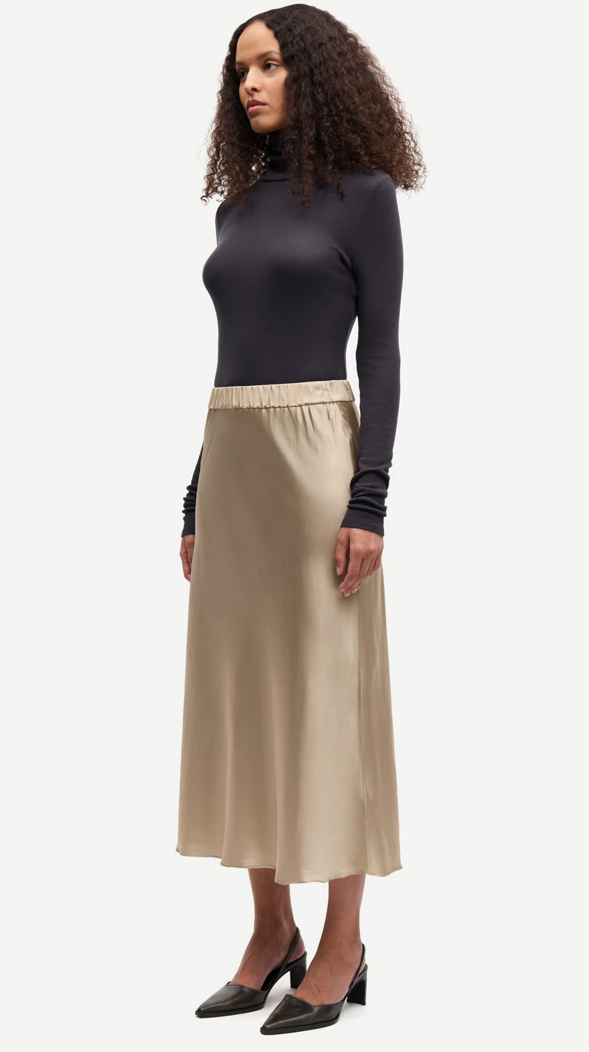 A woman wears the Samsøe Samsøe Samiley Skirt in Aluminium, paired with a fitted black long-sleeve top and black heels, standing against a plain white background.