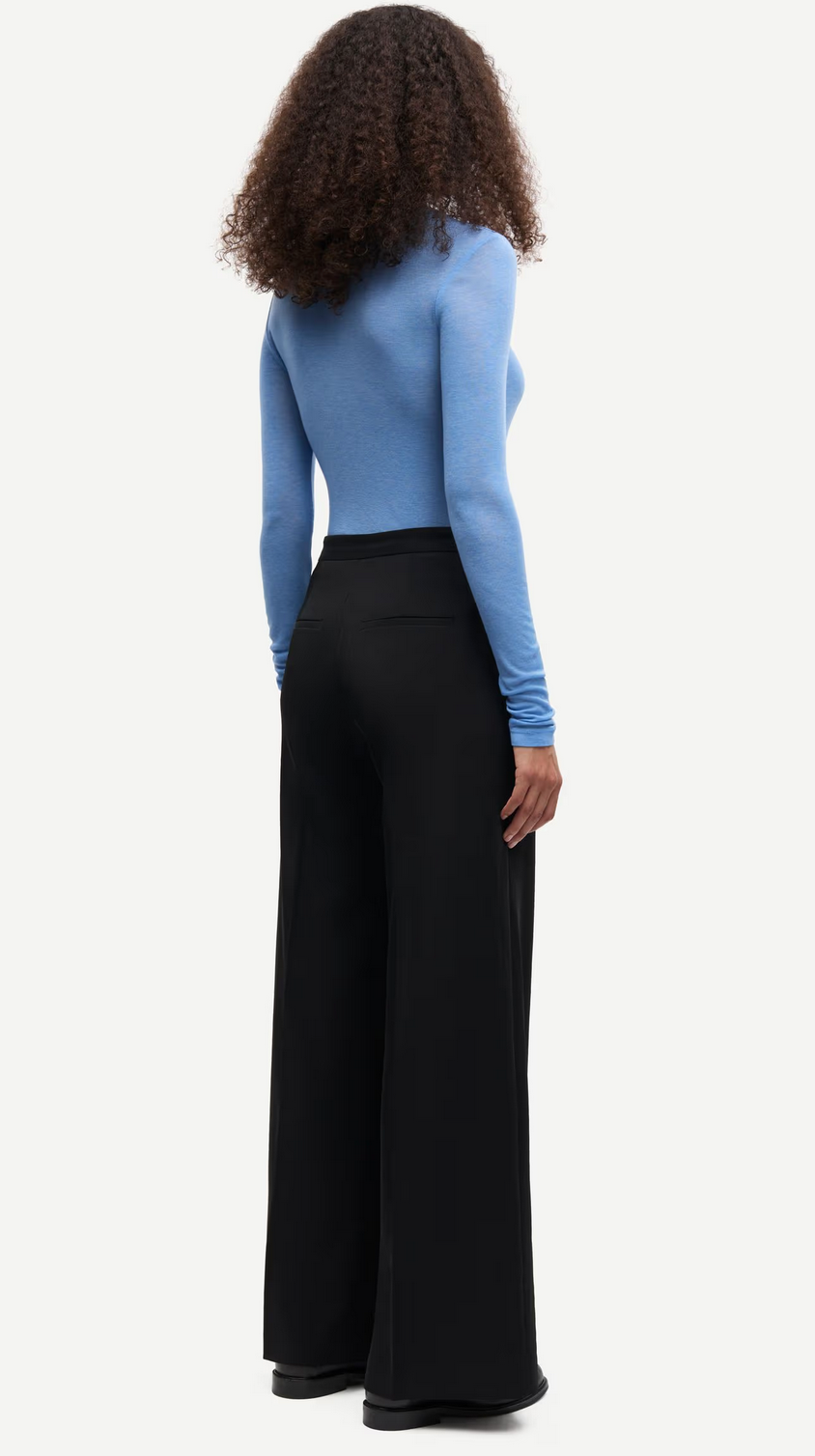 A person stands facing away, wearing the Samsøe Samsøe Sadou T-shirt Long Sleeve in Della Robbia Blue and high-waisted black wide-leg pants with black shoes.