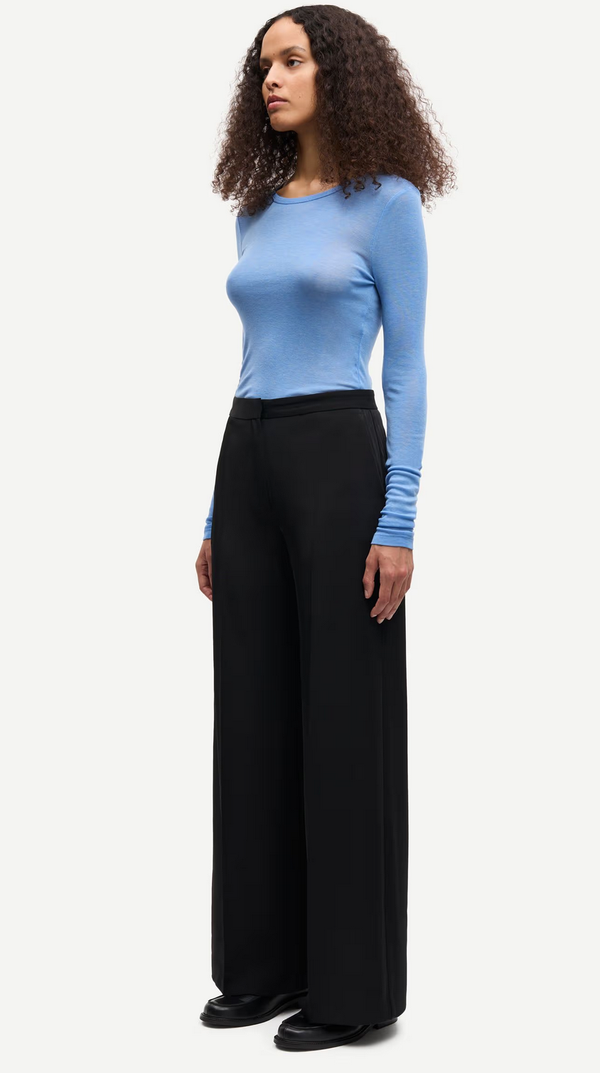 A woman wears the Samsøe Samsøe Sadou T-shirt Long Sleeve in Della Robbia Blue with high-waisted wide-leg black trousers and black shoes, standing against a plain white background.